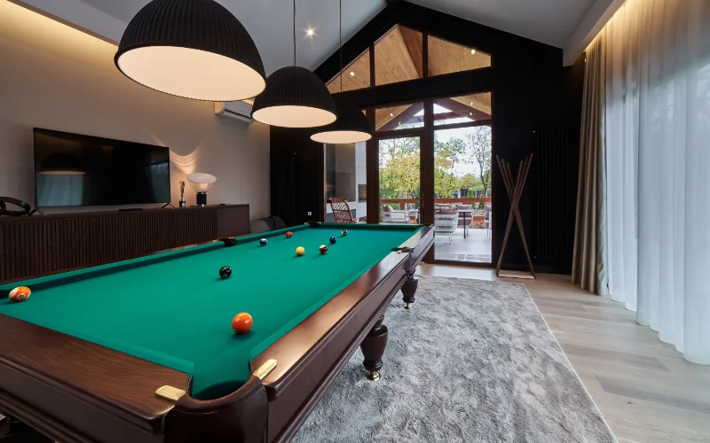 A large green felt pool table centered in a modern game room with vaulted ceilings and dome pendant lights.